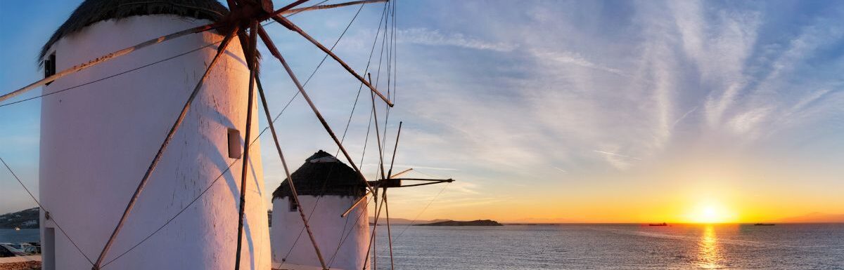 Panorama of traditional Greek windmills on Mykonos island at sunset with dramatic sky and Little Venice quarter with the tourist crowd in Mykonos, Greece in May.