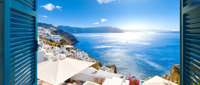 View from an open window with blue shutters of the Aegean sea, caldera, coastline and whitewashed town of Oia, Santorini, Greece.