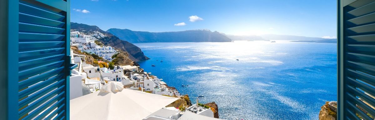 View from an open window with blue shutters of the Aegean sea, caldera, coastline and whitewashed town of Oia, Santorini, Greece.